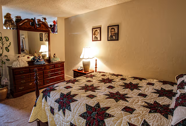 Cozy bedroom with a patterned quilted bed, wooden dresser with mirror, lamp on a nightstand, and framed photos on the wall.