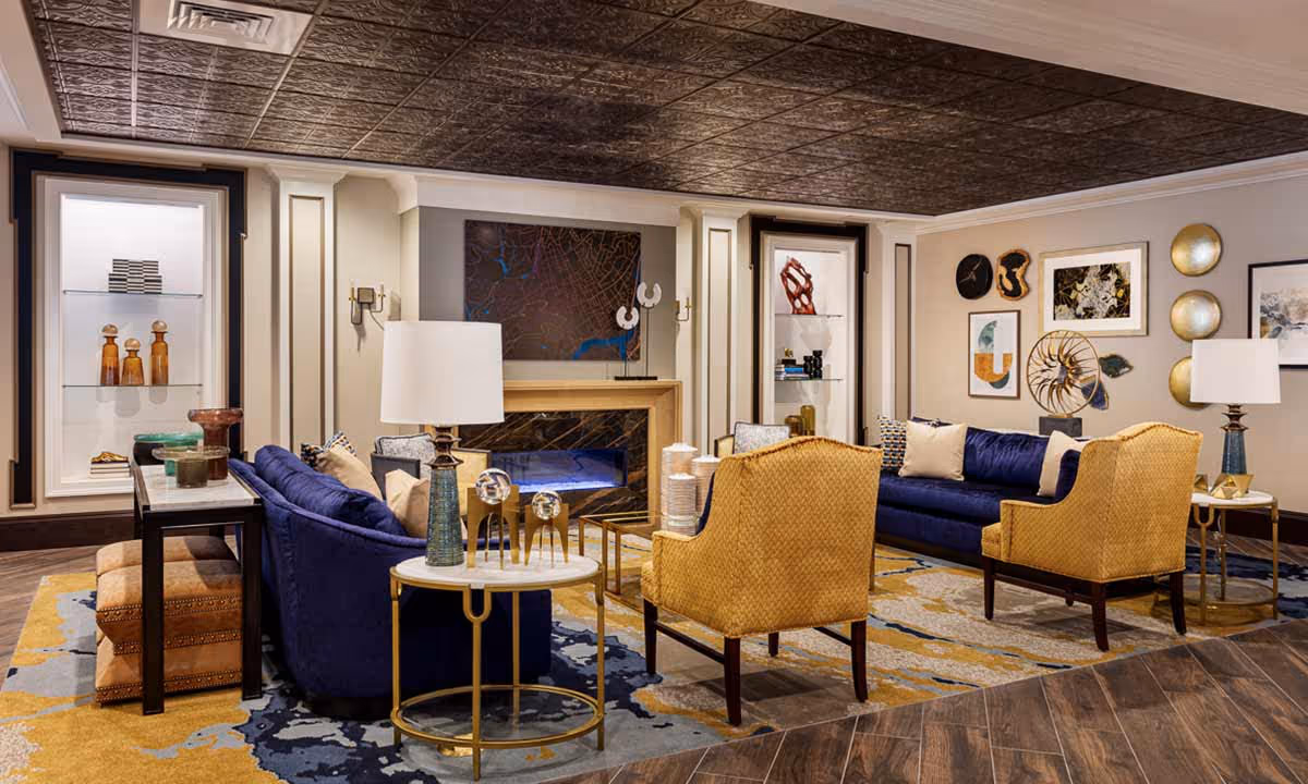 A stylish living room with two blue velvet sofas and two mustard yellow armchairs arranged around a modern fireplace. The room features decorative wall art, built-in shelves with sculptures and books, two side tables with lamps, and a patterned area rug on a wooden floor. The ceiling has a textured dark panel design.