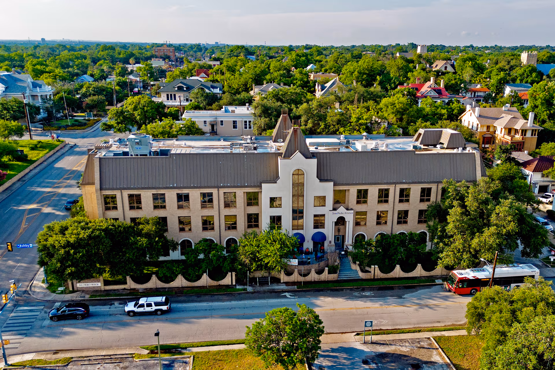 Aerial view of San Pedro Manor, a large three-story building with a gray roof and beige exterior, surrounded by trees and residential houses in a green neighborhood. Several cars and a bus are visible on the streets around the building.