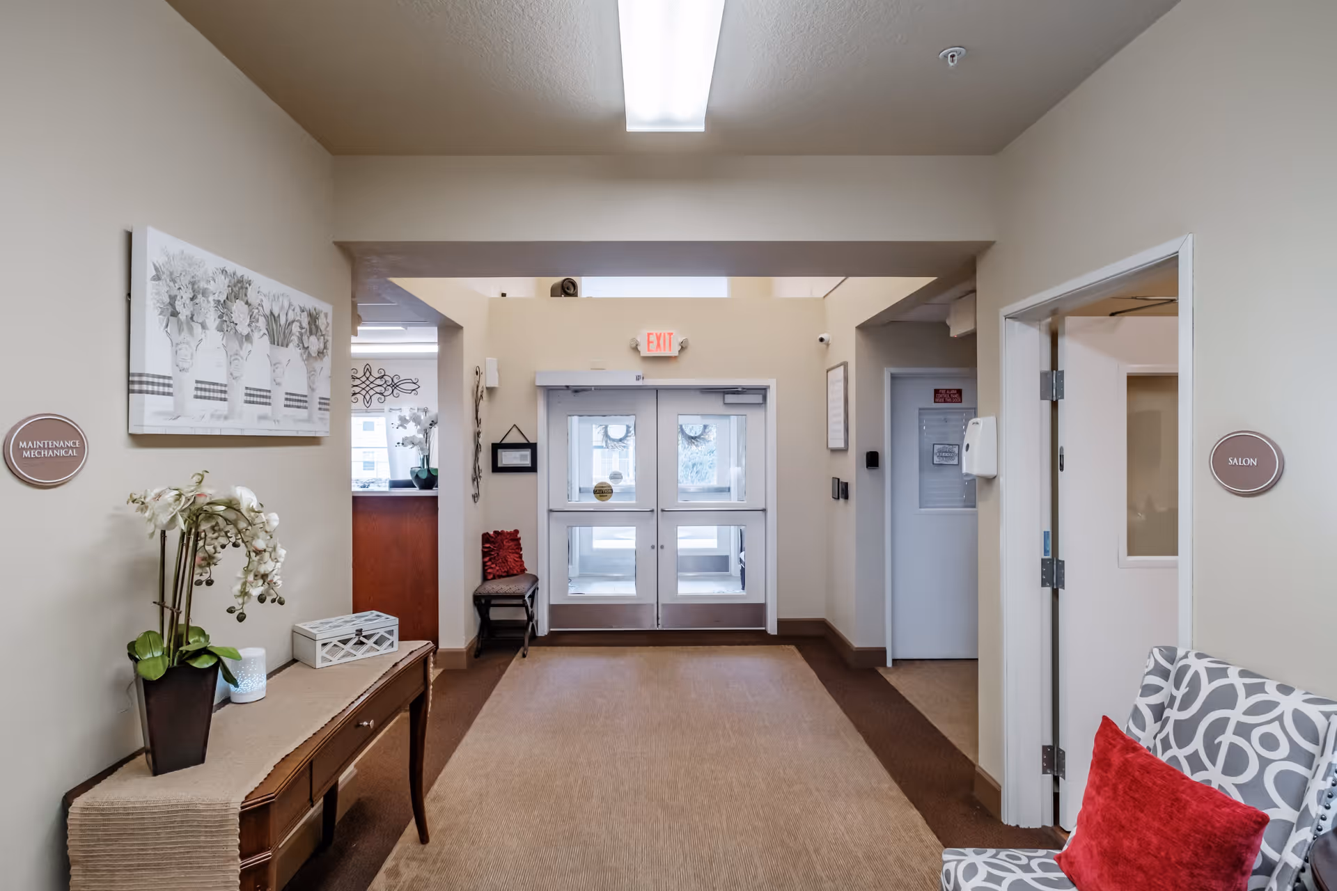 Interior hallway of a senior living facility with double glass exit doors at the end. On the left wall, there is a table with a potted orchid plant and decorative items, and a sign labeled 'Maintenance Mechanical.' On the right wall, there is a chair with a red pillow and a sign labeled 'Salon.' The walls are painted beige, and the floor is carpeted with a brown and tan pattern.
