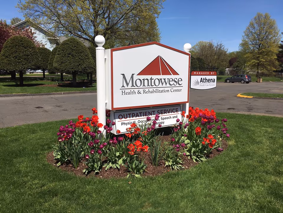 Outdoor view of a sign for Montowese Health & Rehabilitation Center surrounded by a flower bed with red and purple flowers, with trimmed trees and a parking lot in the background under a clear blue sky.