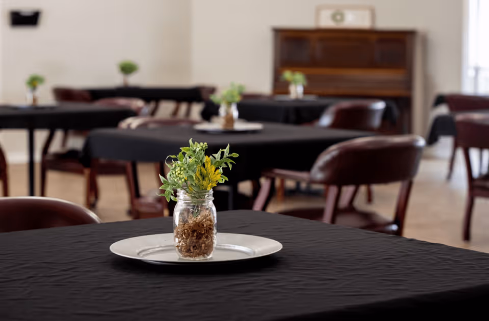 A dining room with multiple tables covered in black tablecloths, each table featuring a small glass jar with a floral arrangement. The room has wooden chairs with dark cushions and a piano against the far wall.