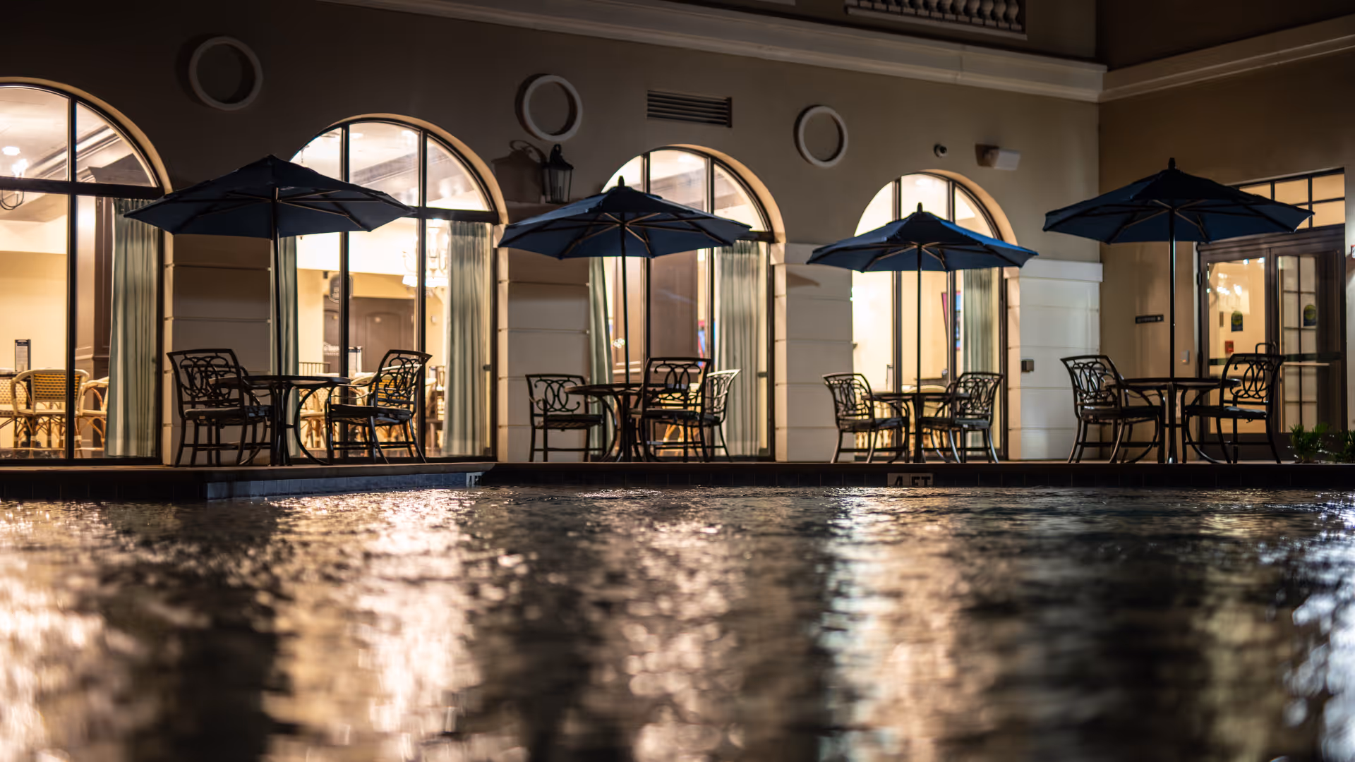 Night view of an outdoor pool area with several tables and chairs under large umbrellas. The background shows a building with large arched windows and warm interior lighting.