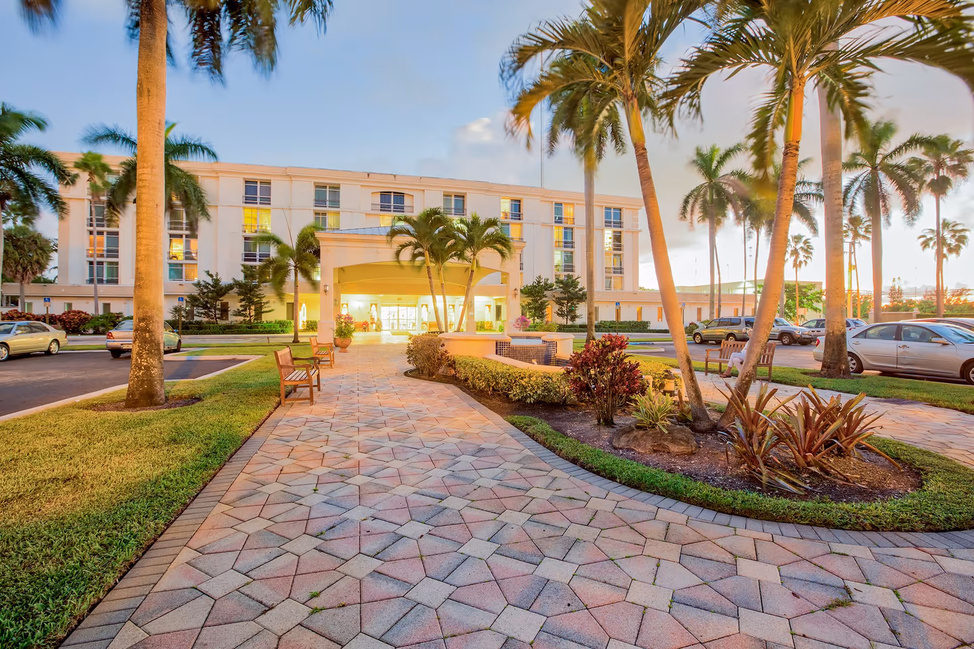 Exterior view of The Peninsula Assisted Living & Memory Care building at sunset, showing a paved walkway lined with benches and palm trees leading to the main entrance. Cars are parked on either side and the building is illuminated with warm lights.