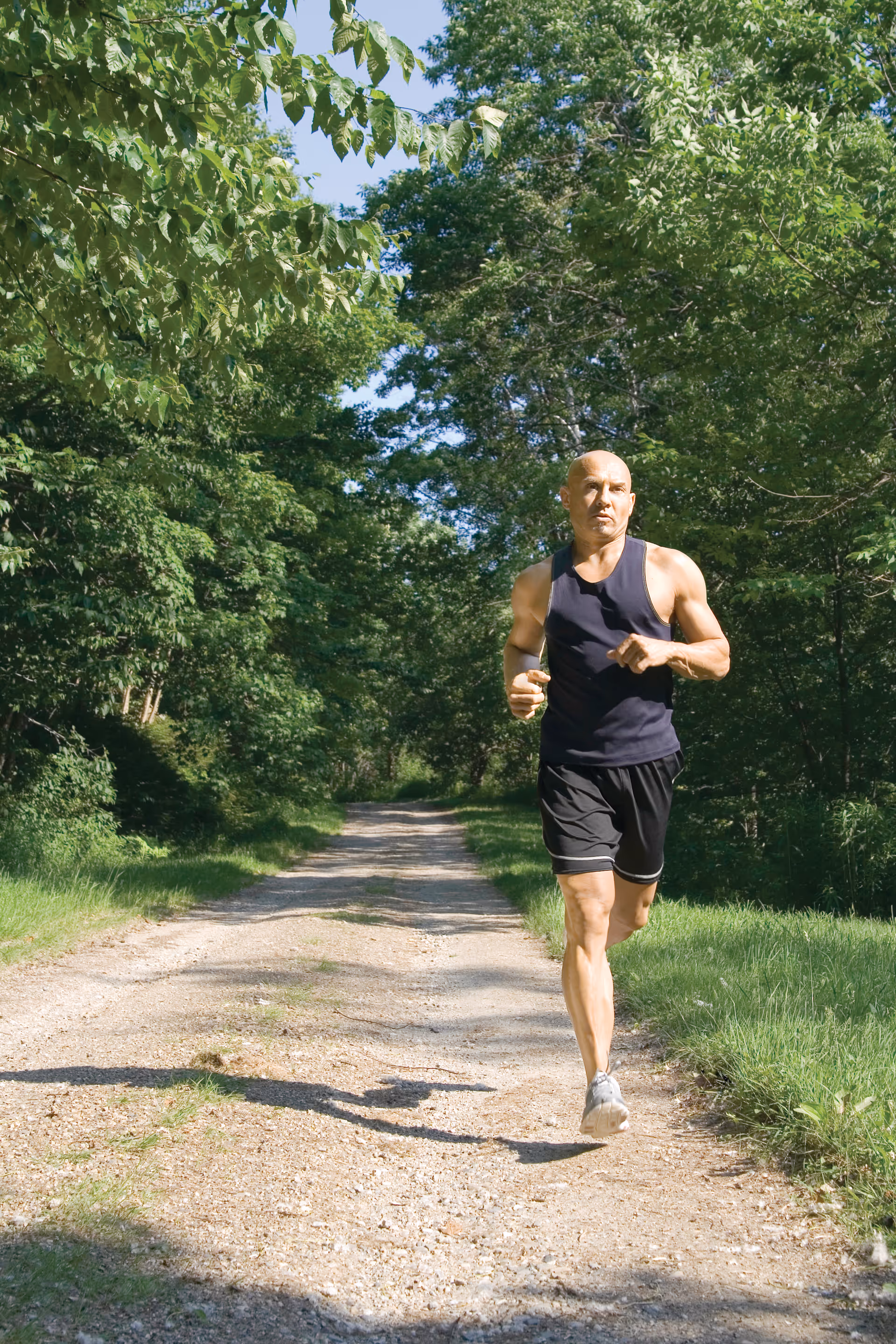 A man wearing a black tank top and black shorts jogging on a gravel path surrounded by green trees and grass on a sunny day.