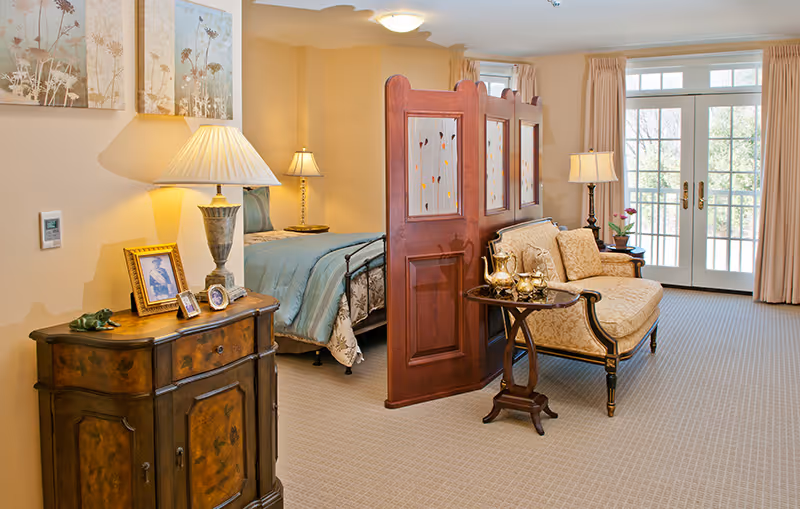 A cozy senior living suite featuring a bed partially separated from a sitting area by a wooden room divider. The sitting area includes an upholstered loveseat and a small round table with a tea set. The room is warmly lit with table lamps and has large French doors with curtains leading outside. A wooden cabinet with framed photos and a lamp is also visible.