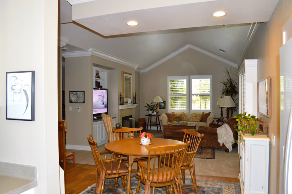 View of a cozy living and dining area in a senior living facility. The dining area features a round wooden table with four matching chairs and a small flower centerpiece. The living room has a beige sofa, a brown armchair, a wooden coffee table, and a TV mounted above a fireplace. Large windows with white shutters allow natural light to fill the room. The walls are painted in neutral tones, and there are various plants and framed artwork decorating the space.