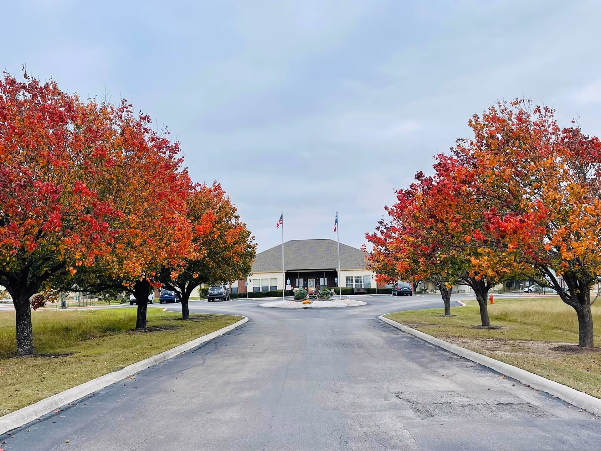 A paved driveway lined with trees displaying vibrant red and orange autumn foliage leads to a single-story building with a gray roof. Two flagpoles with flags stand in front of the building, and a few cars are parked near the entrance under an overcast sky.