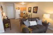 Open living room and dining area in a senior apartment with a sofa, dining table, and kitchen in the background.