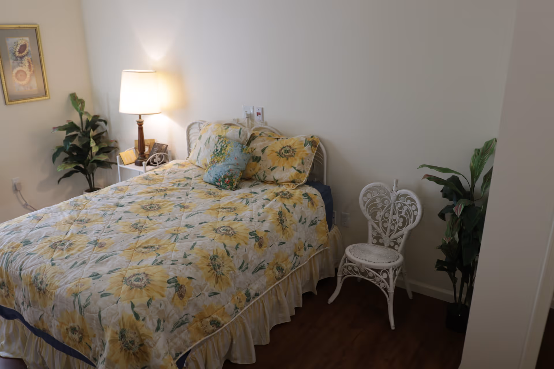 A small bedroom with a sunflower-patterned bed, lit bedside lamp, white decorative chair, and potted plants.