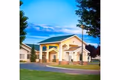 Front entrance of a single-story assisted living building with columns, a circular driveway, landscaped lawn and trees under a blue sky.