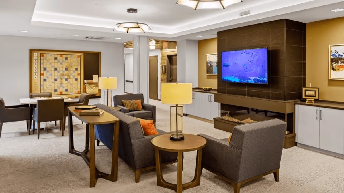 A cozy living room area in a memory care facility featuring gray armchairs and a sofa with orange and brown accent pillows, a round wooden side table with a lamp, a wall-mounted flat-screen TV above a modern fireplace, and a large Scrabble game board on the wall near a table with chairs for games or activities.