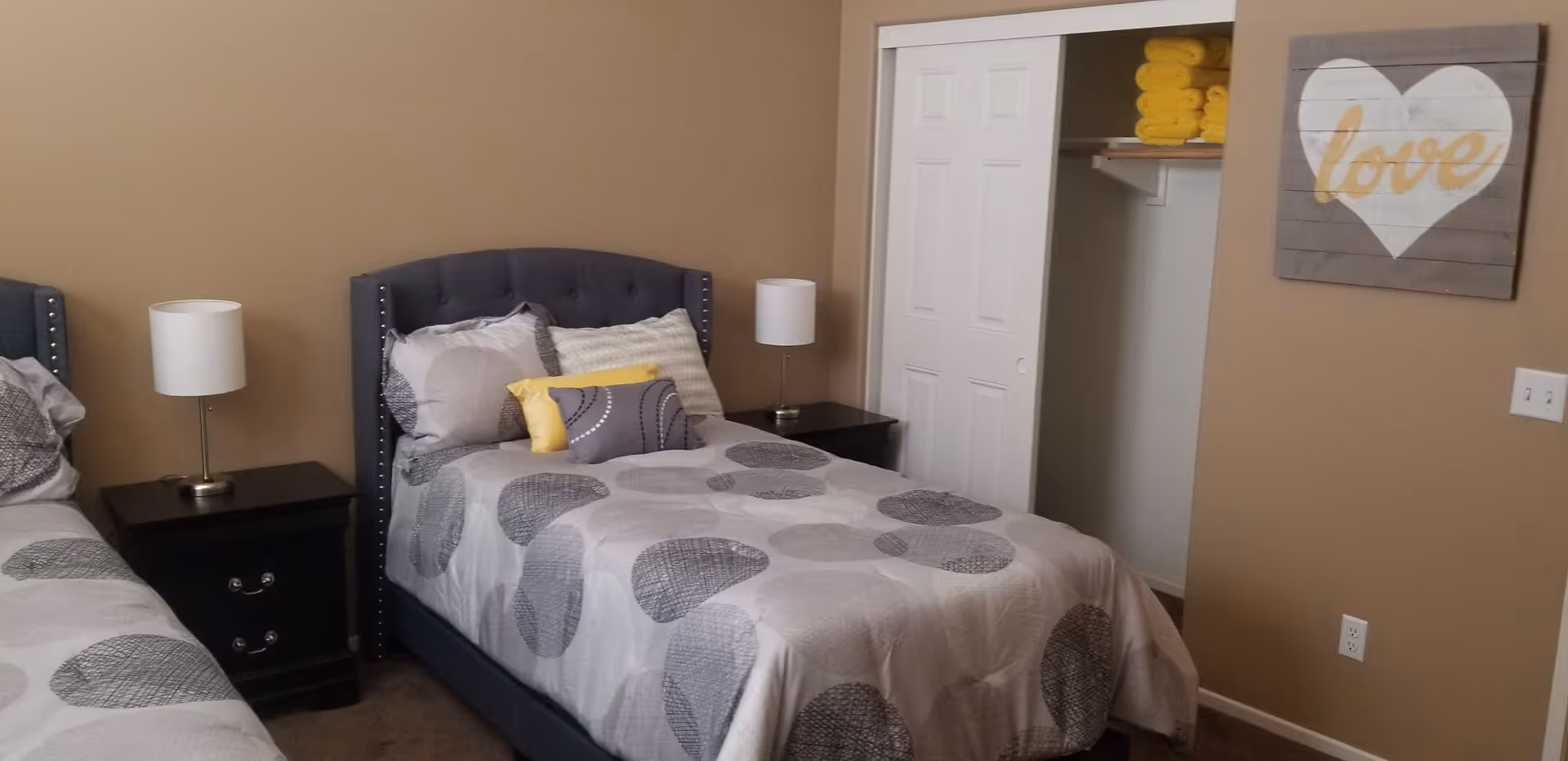 Two single beds in a beige bedroom with gray patterned bedding, nightstands and lamps, an open closet with yellow towels, and a wall sign that reads 'love'.