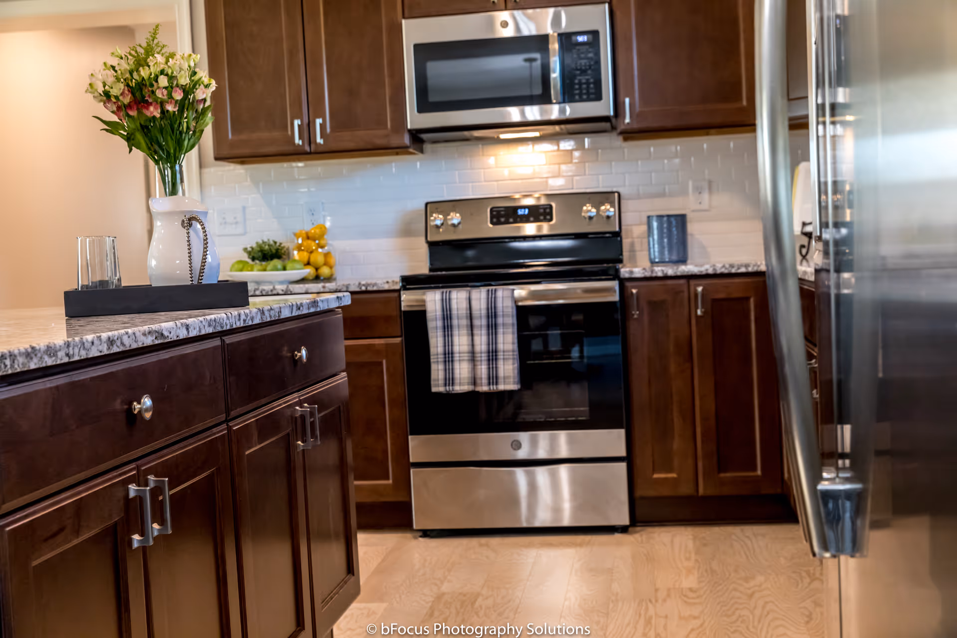 Modern kitchen with dark wood cabinets, stainless steel appliances including a stove with a plaid towel hanging on the handle, a microwave above the stove, and a refrigerator on the right. A countertop with a vase of flowers and a glass is visible in the foreground.