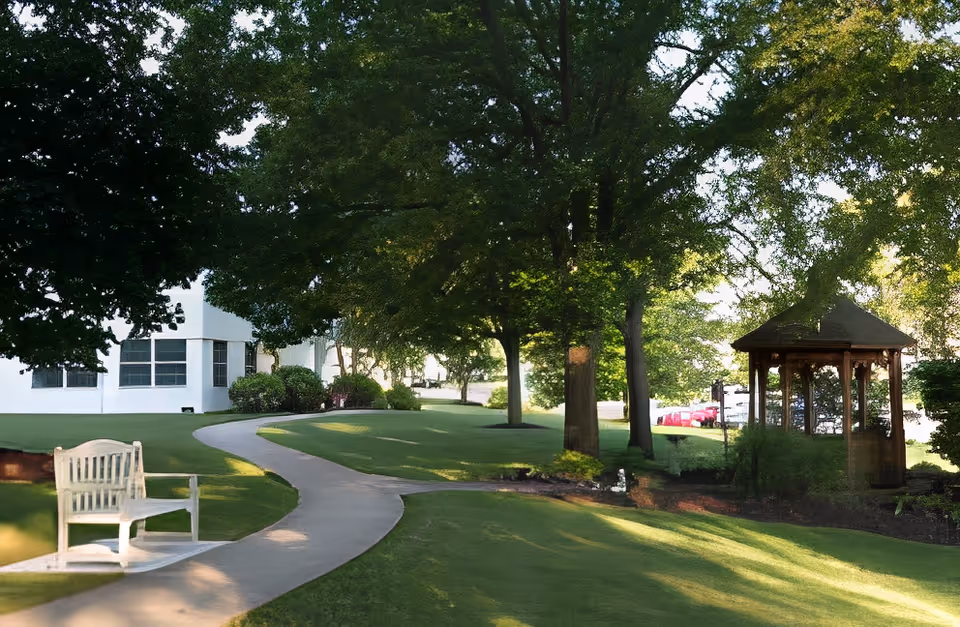 A peaceful outdoor garden area with a winding concrete pathway, a wooden bench on the left, large green trees providing shade, and a wooden gazebo on the right. A white building is partially visible in the background.