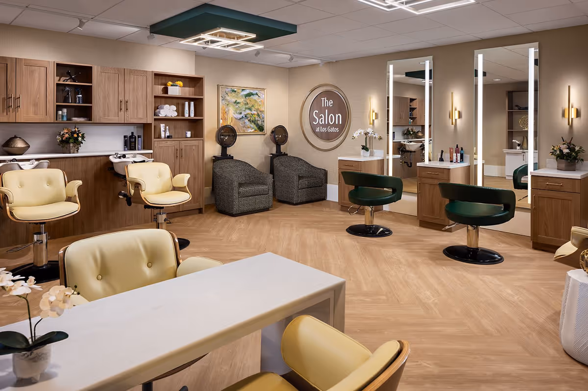 Interior view of a senior living facility salon with beige and green styling chairs, large mirrors with lights, wooden cabinets, hair washing stations, and a sign on the wall that reads 'The Salon at Los Gatos'. The room has a warm, inviting atmosphere with wood flooring and modern lighting fixtures.