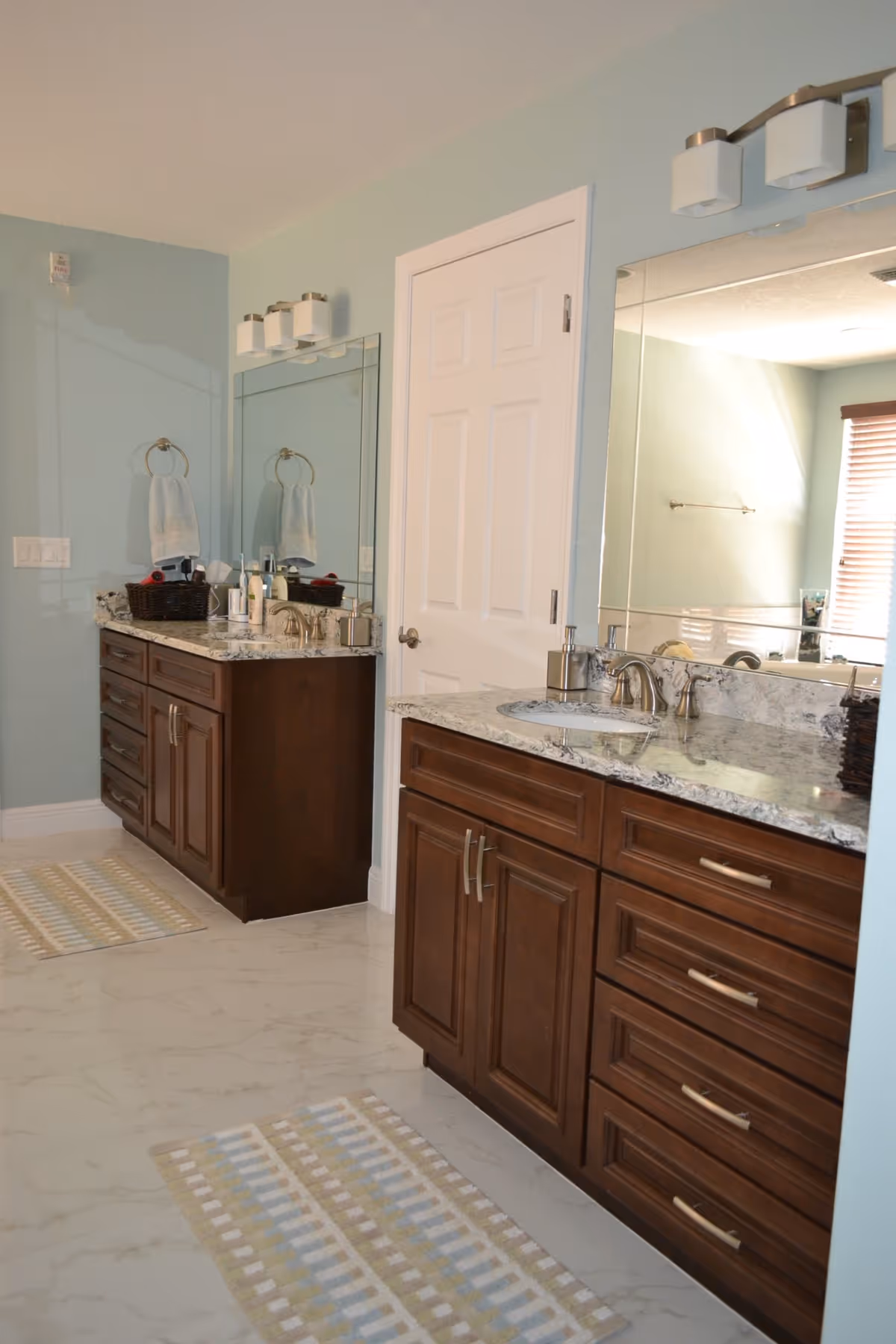 Bright bathroom with two marble-topped vanities, dark wood cabinets, large mirrors, and floor rugs.