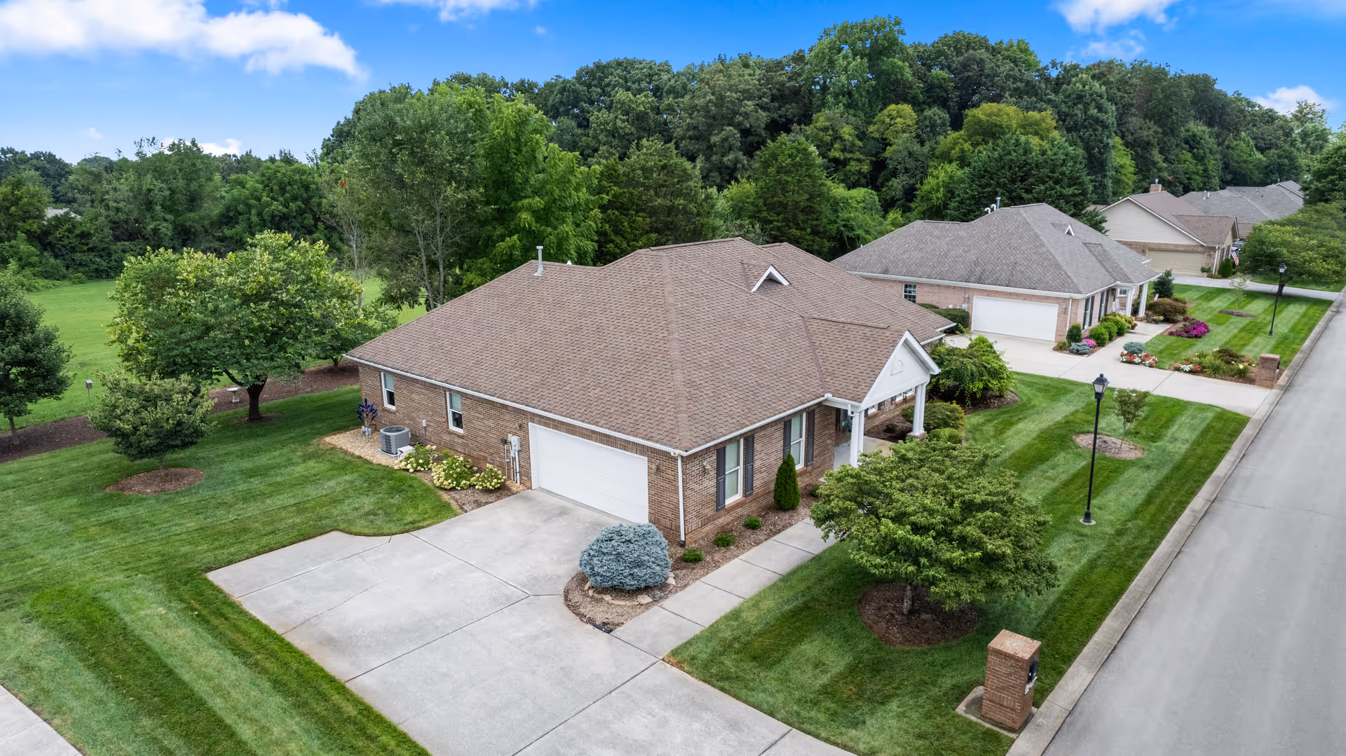 Aerial view of a single-story brick residential building with a brown shingled roof, attached garage, and well-maintained green lawn with trees and shrubs. The building is part of a neighborhood with similar houses and a paved street running alongside.
