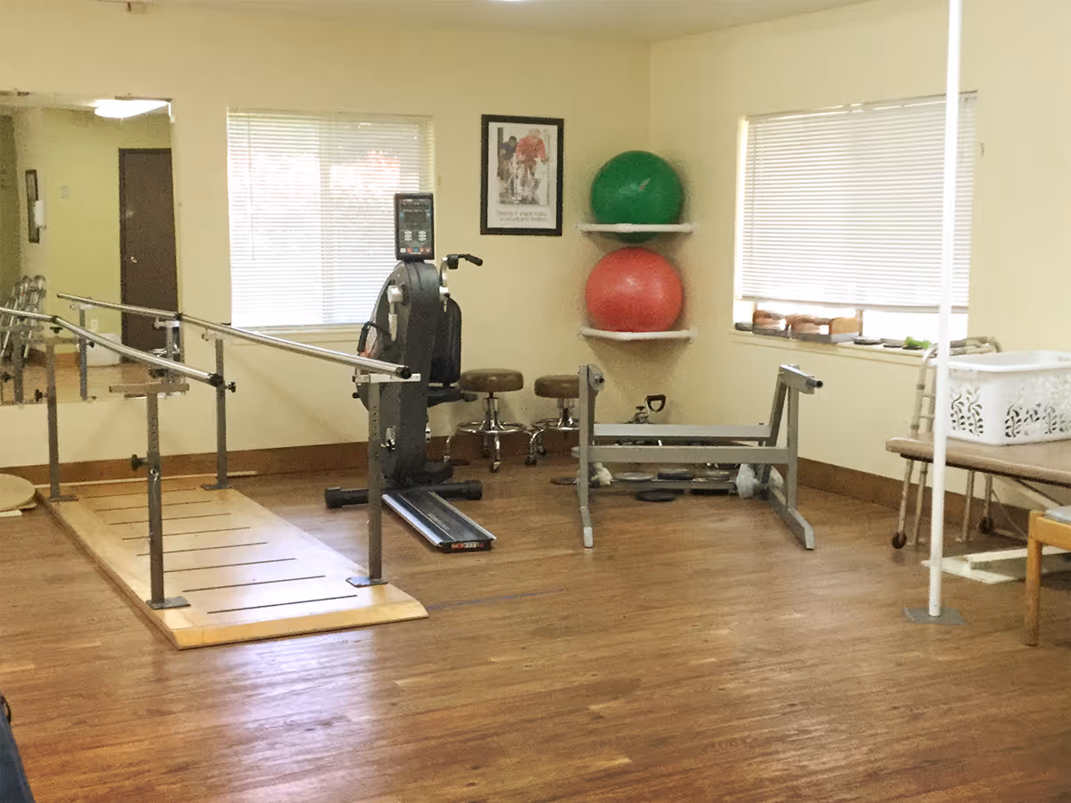 A rehabilitation room with parallel bars for walking exercises, a rowing machine, two exercise balls on wall shelves, stools, and various physical therapy equipment. The room has wooden flooring, two windows with blinds, and light-colored walls.