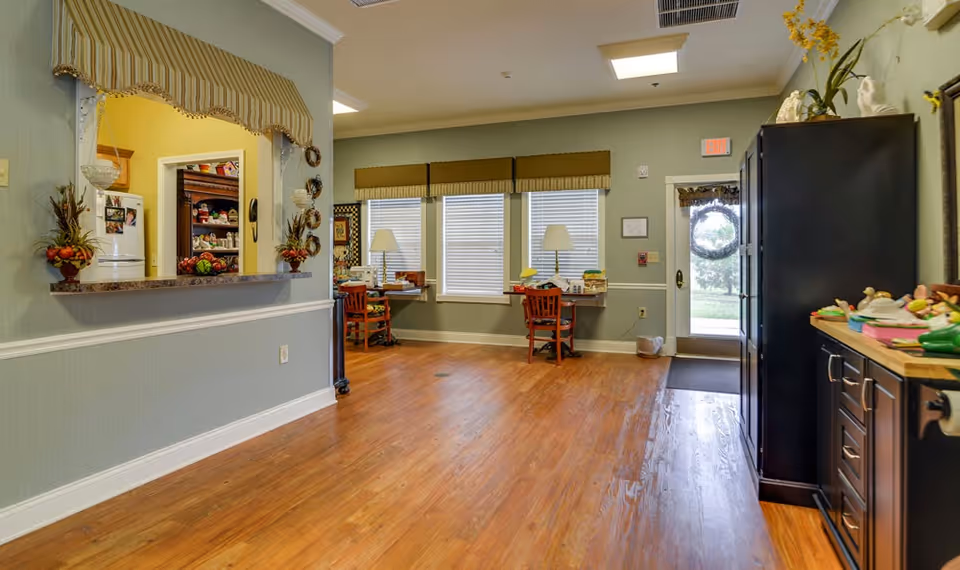 Interior view of a senior living facility room with wooden flooring, two desks with chairs and lamps near three windows with blinds, a black cabinet on the right, and a pass-through window with a striped valance showing a kitchen area with a refrigerator.