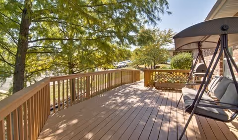 A spacious wooden deck attached to a building, featuring a cushioned swing seat on the right side. The deck overlooks a green area with trees and a parking lot in the background under a sunny sky.