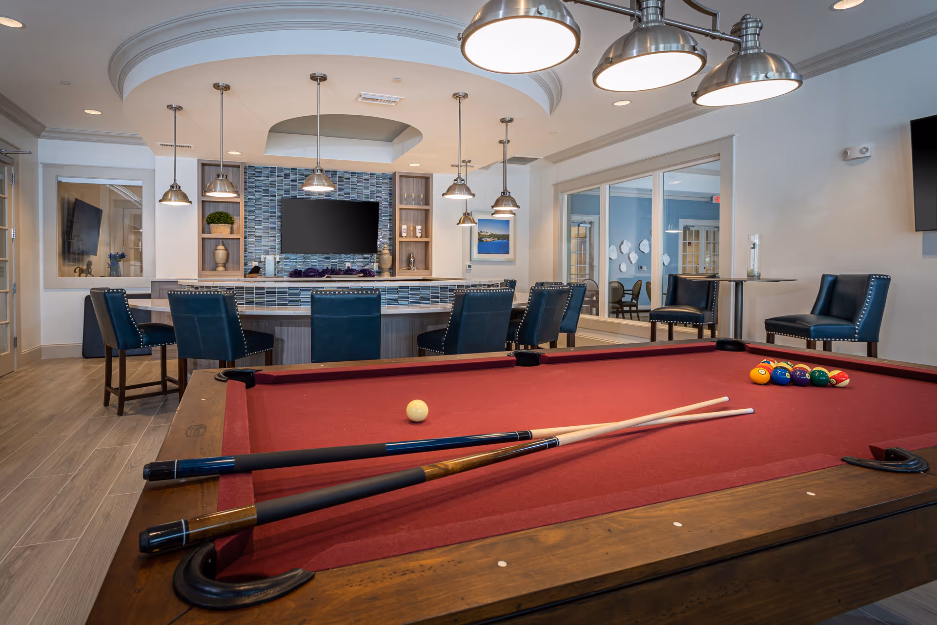 Interior view of a recreational room in a senior living facility featuring a red felt pool table with two cues and billiard balls arranged for a game. Behind the pool table is a bar area with high chairs, pendant lights, a TV mounted on a tiled wall, and decorative shelves. The room has large windows and additional seating with blue upholstered chairs.