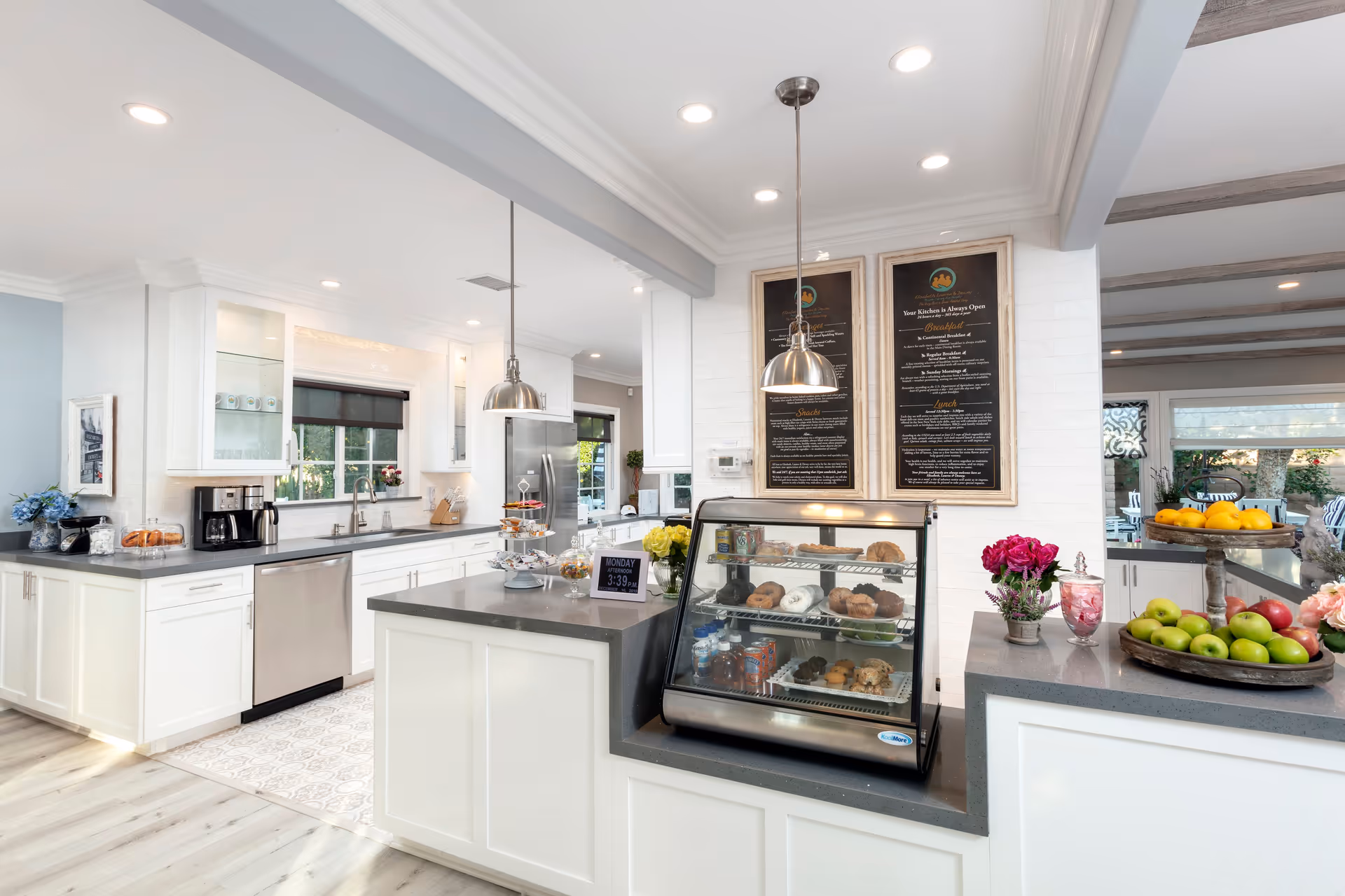 Bright and modern kitchen area with white cabinetry and gray countertops. There is a display case with assorted pastries and beverages on the counter, two hanging pendant lights, and framed menu boards on the wall. Fresh fruits and flowers are arranged on the counter, and large windows provide natural light.