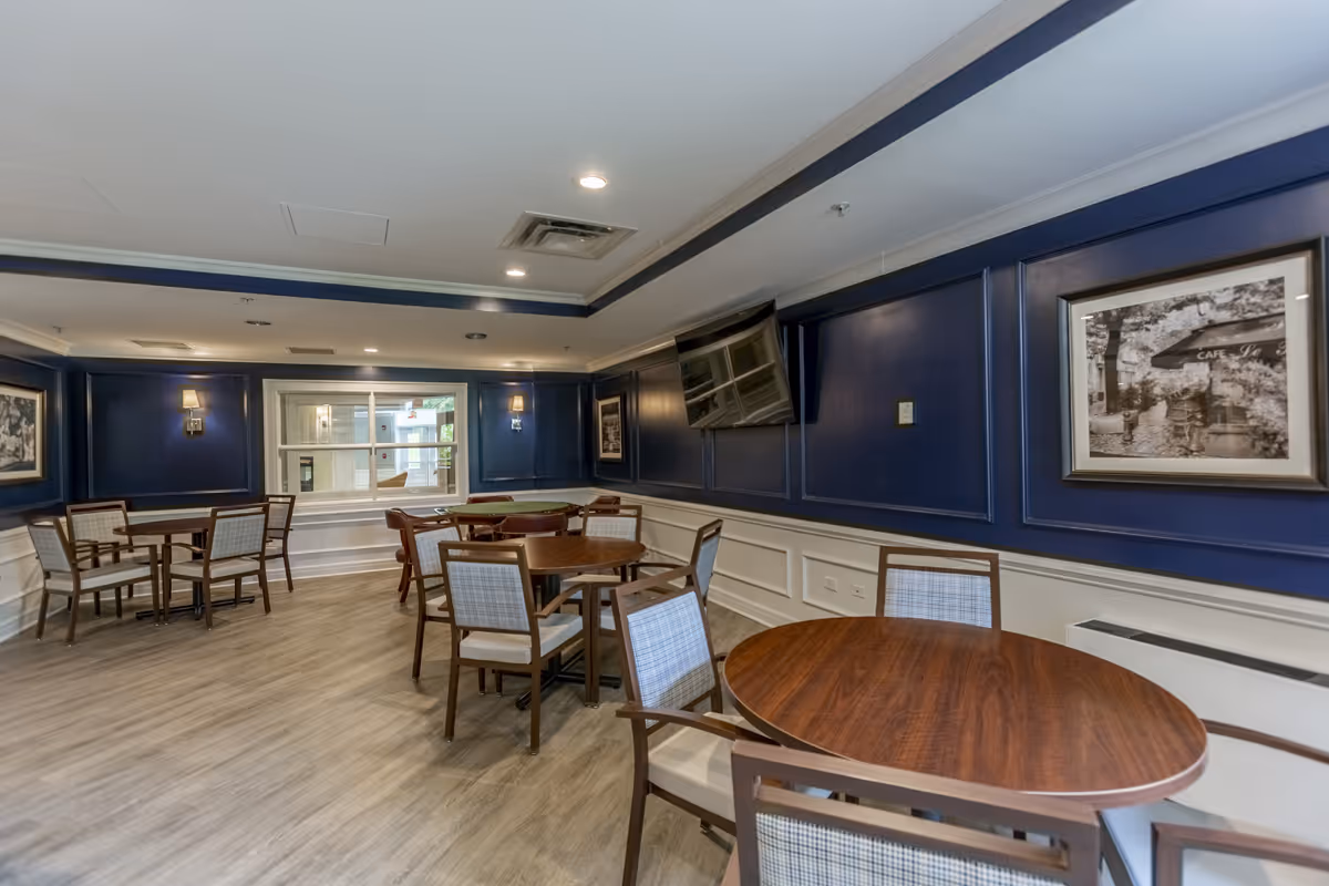A dining-style common room with round wooden tables, chairs, a wall-mounted TV, and dark blue paneled walls.