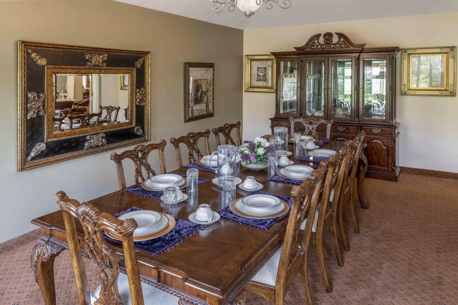 A formal dining room with a long wooden table set for eight people. The table is adorned with white plates, cups, glasses, and purple placemats. A floral centerpiece is placed in the middle. The room features ornate wooden chairs, a large decorative mirror on one wall, framed artwork, and a wooden china cabinet with glass doors displaying dishes.