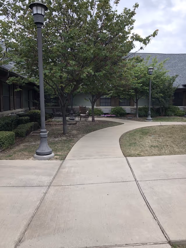 Curved concrete walkway through a courtyard with trees, lampposts, and seating in front of a single-story building.