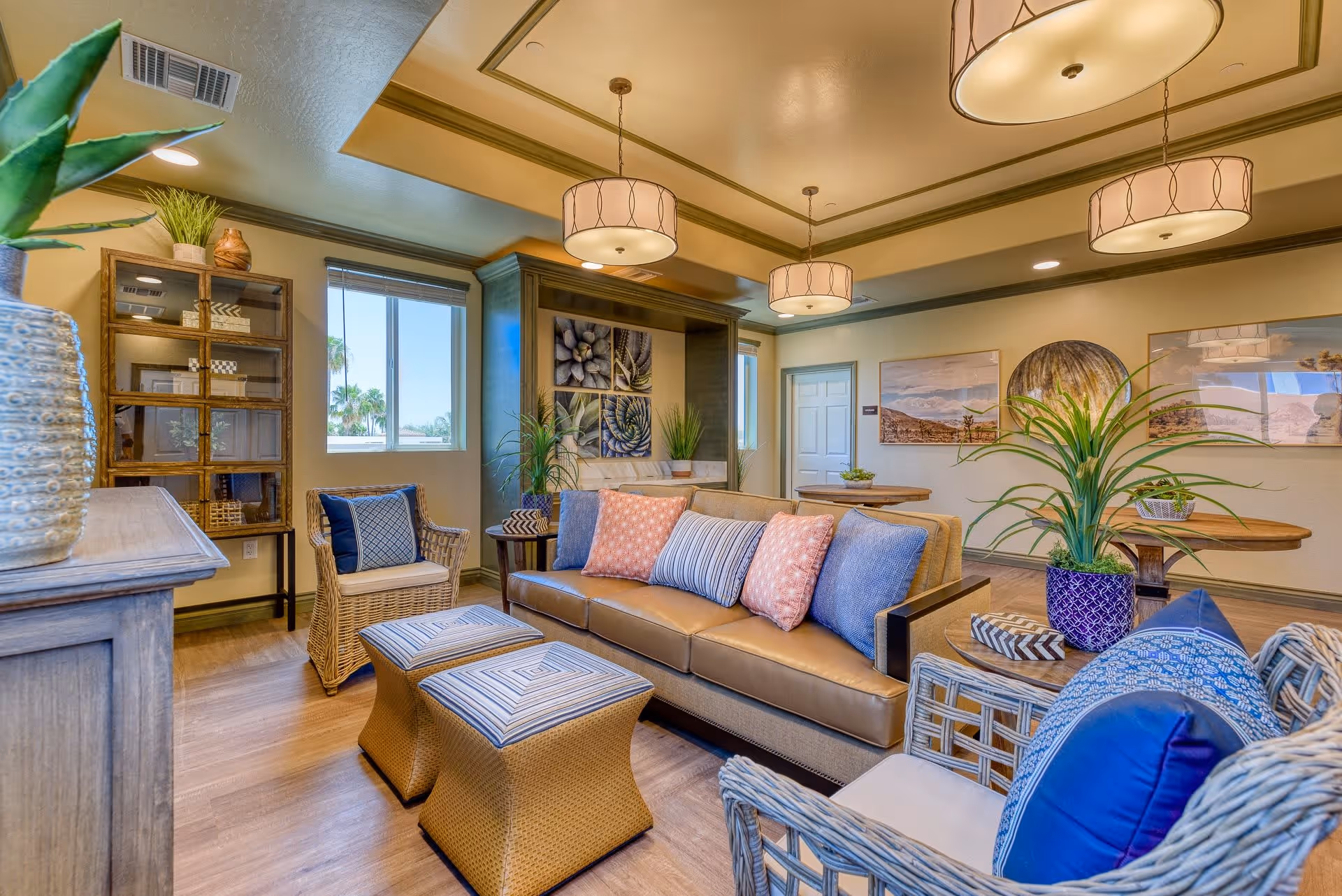 A bright and inviting living room area with a tan leather sofa adorned with blue and pink pillows, two wicker chairs with blue cushions, and two wicker ottomans with striped cushions. The room features wooden flooring, a wooden cabinet with glass doors, potted plants, framed artwork on the walls, and three pendant lights hanging from the ceiling. Windows allow natural light to fill the space.