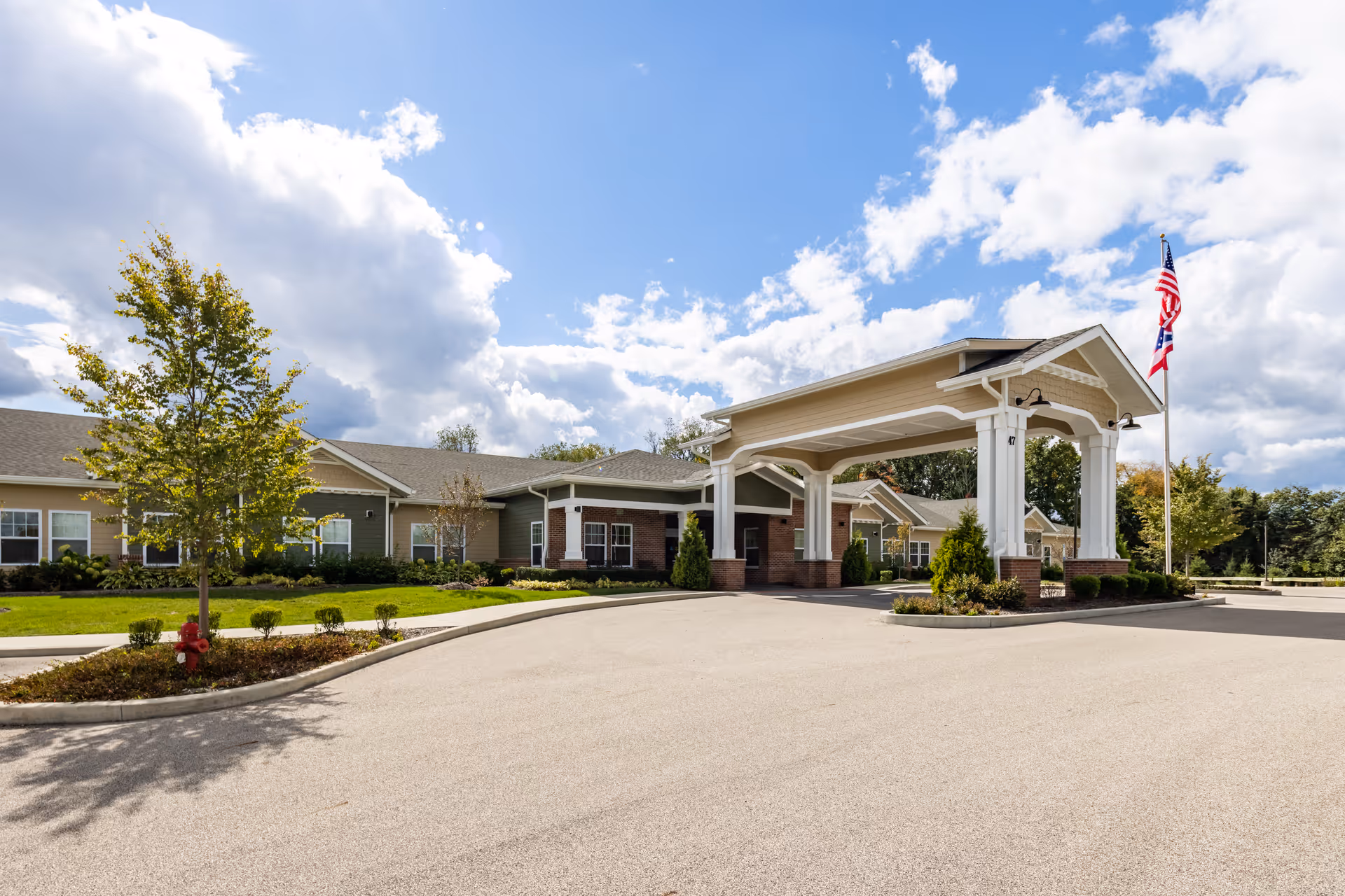 Front entrance of a memory care building with a covered porte-cochère, landscaped lawns, and flagpoles under a partly cloudy sky.