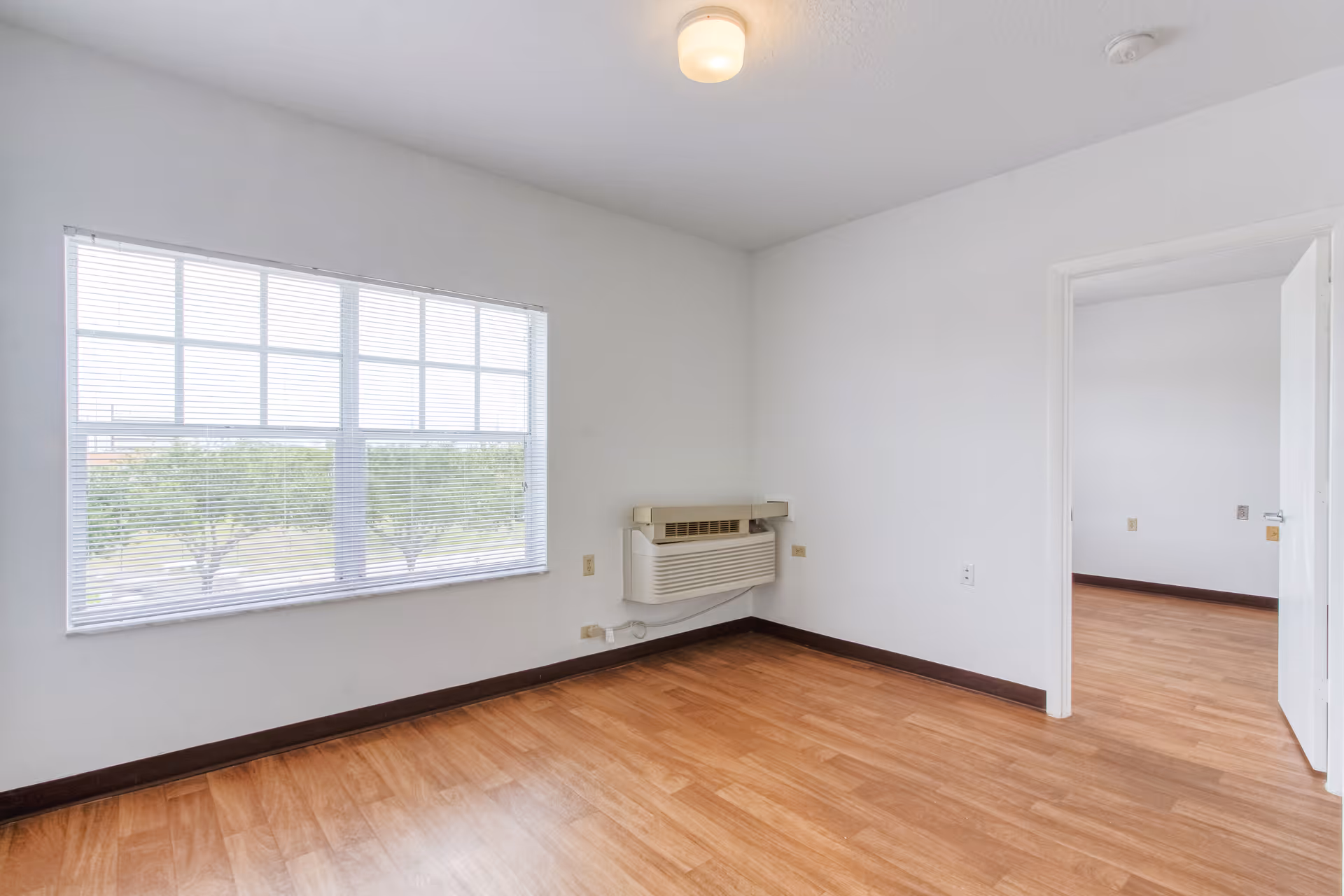 Empty room with light wood flooring, white walls, a large window with blinds, a wall-mounted air conditioning unit, and an open door leading to another similar room.