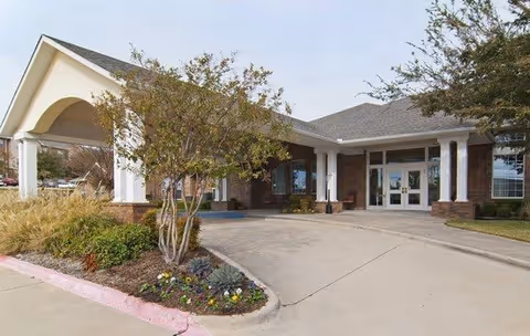 Front exterior view of Morada Cedar Hill Assisted Living and Memory Care facility showing the entrance with a covered driveway, landscaped garden with small trees and flowers, and a brick building with white columns and large windows.