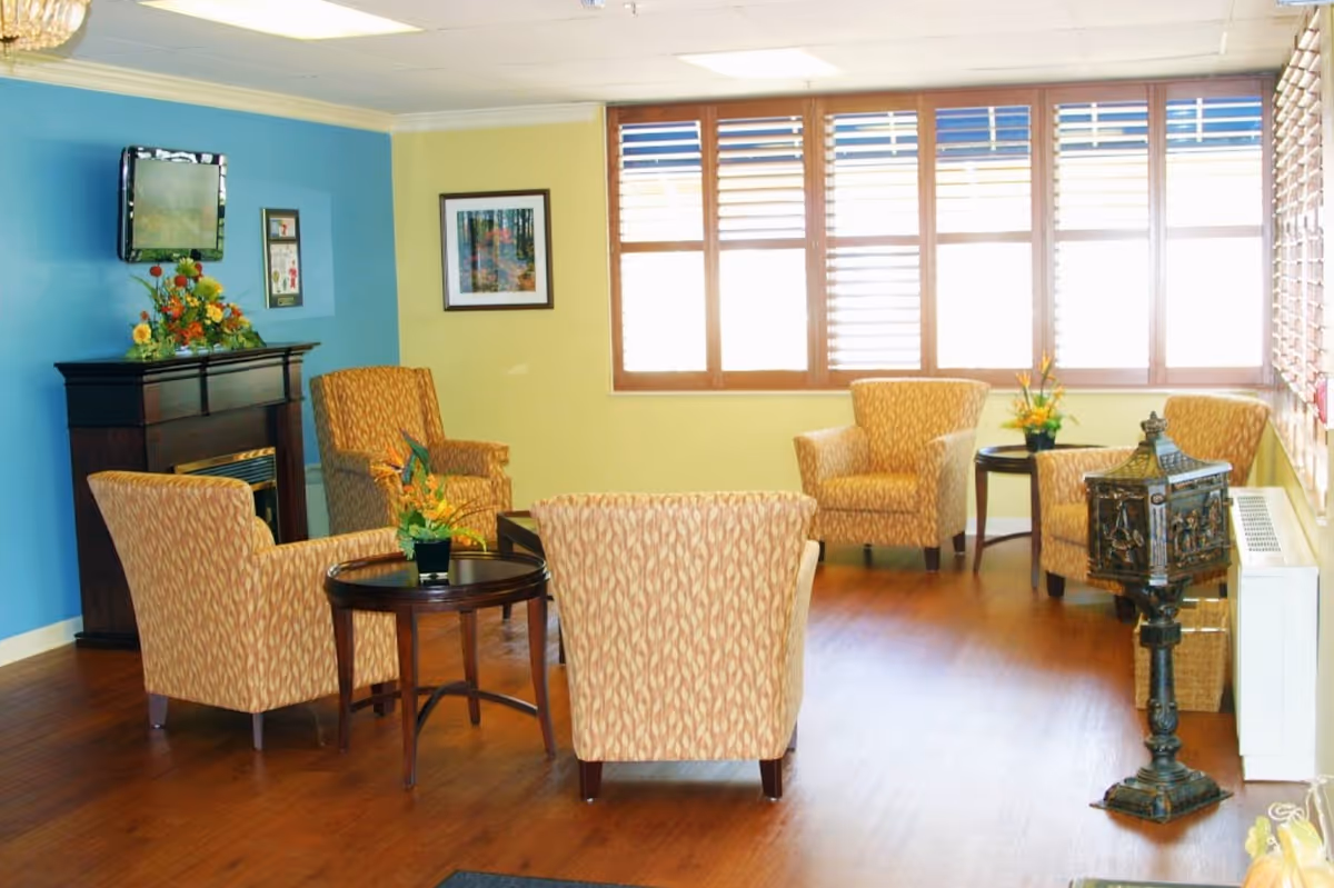 A bright and cozy sitting area with five patterned armchairs arranged around two small round wooden tables with flower arrangements. The room has wooden flooring, a blue and yellow painted wall, a wall-mounted TV above a dark wooden fireplace mantel decorated with flowers, large windows with wooden blinds letting in natural light, and a decorative metal stand near the window.