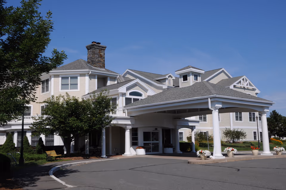 Exterior view of Lathrop Retirement Community building with a covered entrance, white columns, and a stone chimney under a clear blue sky. There are trees, shrubs, and flower pots around the entrance area.