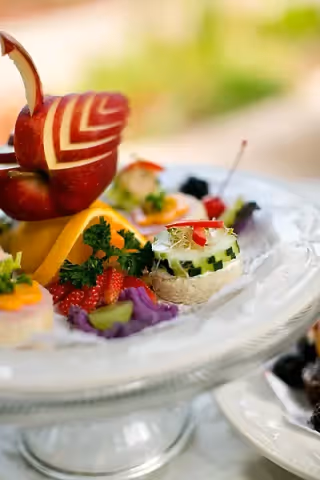 A decorative food platter featuring an intricately carved red apple shaped like a bird, surrounded by various colorful fruits, vegetables, and garnishes on a glass serving dish.