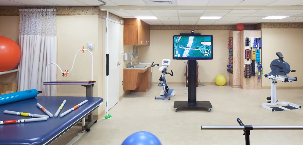 A physical therapy or rehabilitation room with exercise equipment including stationary bikes, therapy balls, resistance bands, and a large screen monitor. The room has beige walls, a blue therapy table with colored markers, and wooden cabinets with a sink.