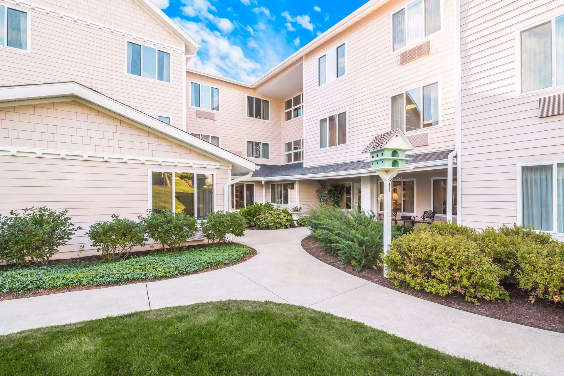 Courtyard and entrance of a light-pink multi-story senior living building with pathways, shrubs, and a decorative birdhouse.