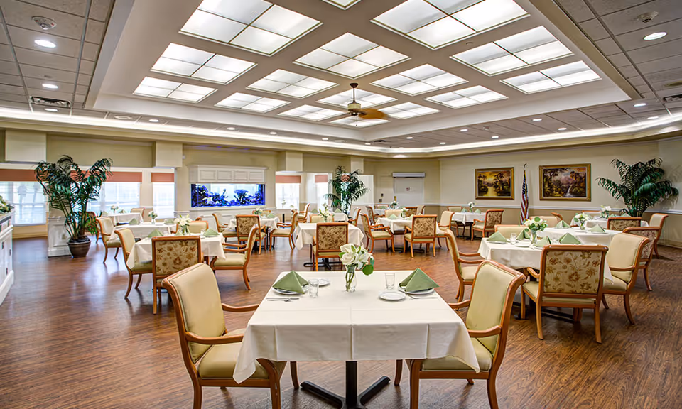 A spacious dining room in a senior living facility with multiple tables covered in white tablecloths, each set with green folded napkins, plates, and glasses. The room features wooden flooring, large windows with blinds, potted plants, framed paintings on the walls, and a ceiling with recessed lighting and a ceiling fan.