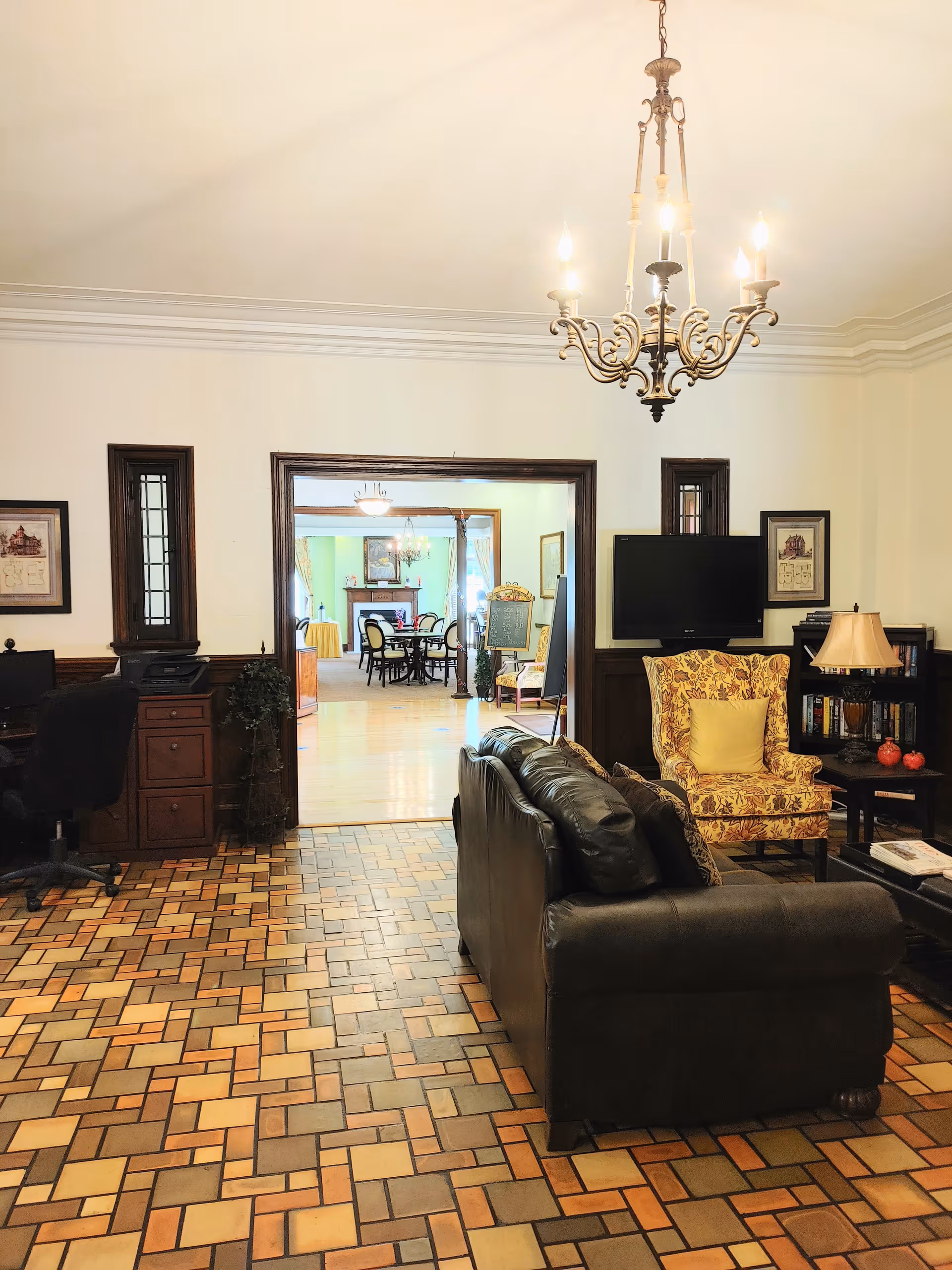 Interior view of a senior living facility lounge area with a black leather sofa, a floral patterned armchair with a yellow cushion, a coffee table, a bookshelf, and a wall-mounted TV. The floor has a patterned tile design, and a chandelier hangs from the ceiling. Through an open doorway, a dining area with tables and chairs is visible.