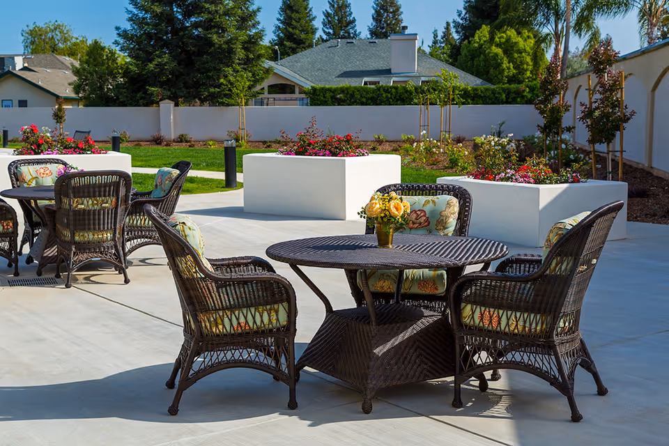 Wicker patio table and chairs with floral cushions on a paved outdoor seating area surrounded by planters and landscaping.