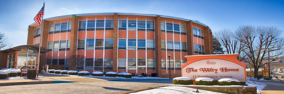 Curved three-story brick senior care building with an American flag and a front sign reading 'The Welty Home' on a snowy lawn.