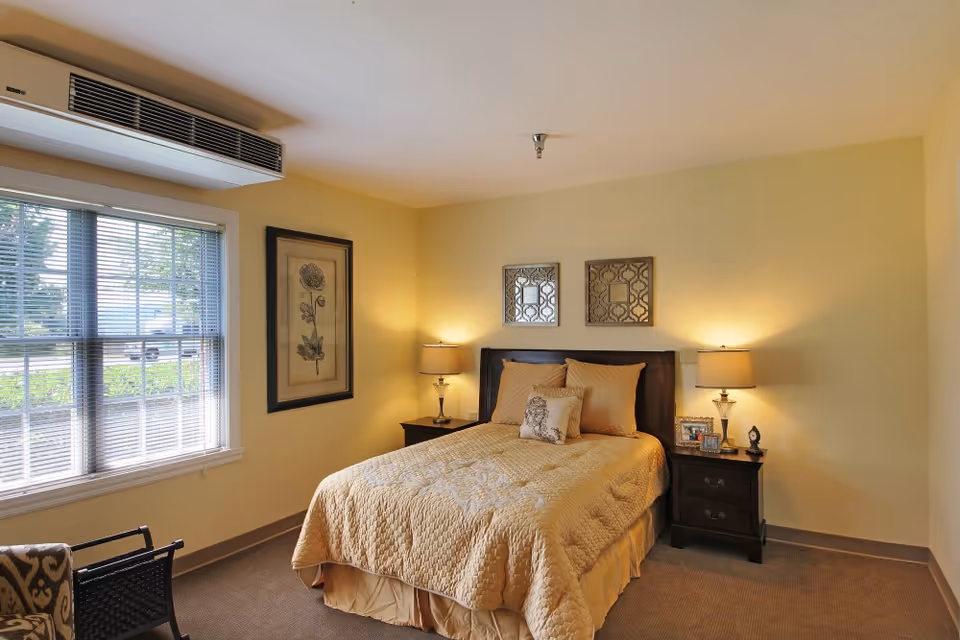 A cozy bedroom with a double bed covered in a beige quilt and matching pillows. There are two bedside tables with lamps on each side of the bed. Above the bed, two decorative mirrors are hung on the wall. A framed botanical print is on the wall to the left, next to a window with blinds partially open, letting in natural light. The room has beige walls and carpeted floor.