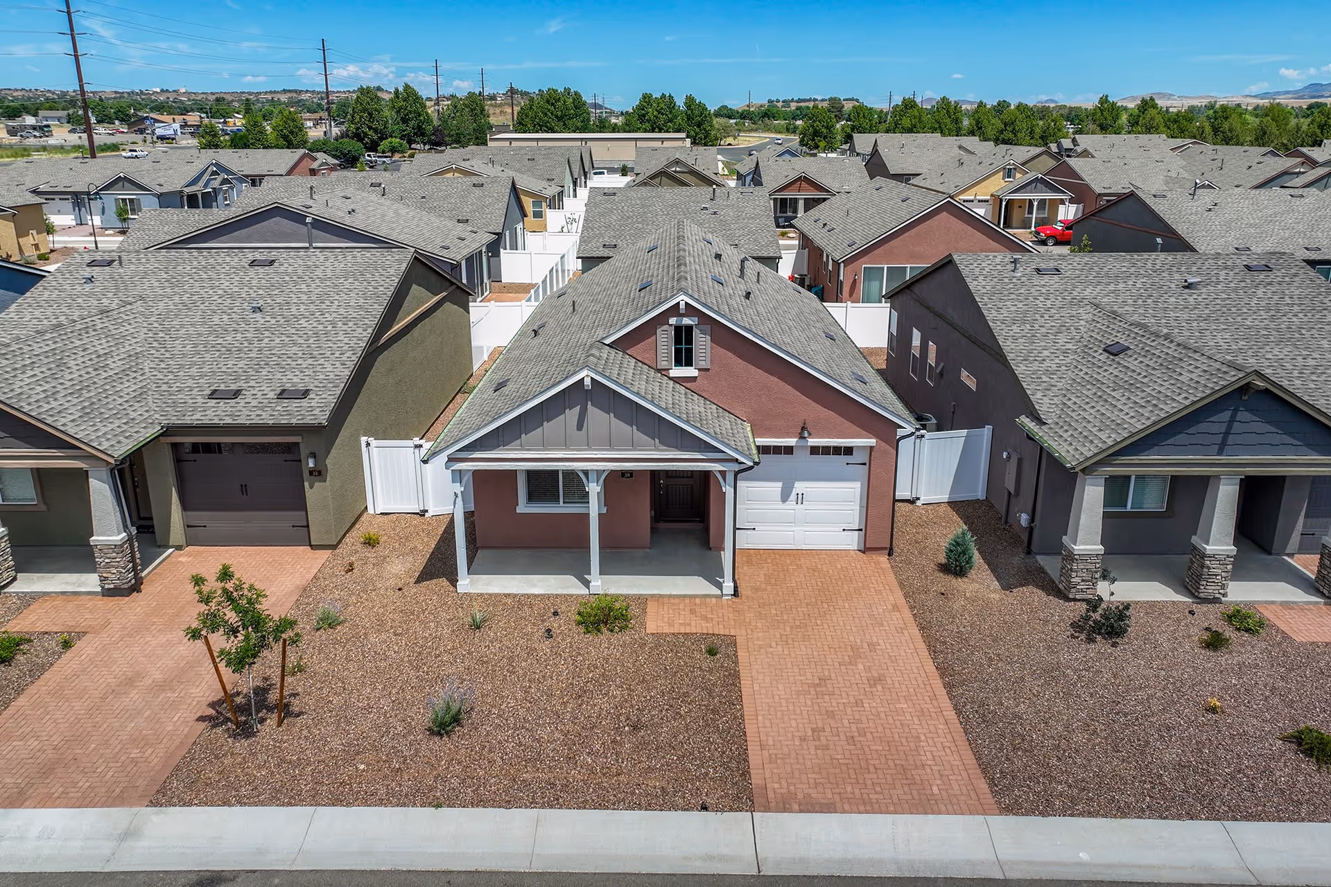 Aerial view of a residential neighborhood showing a row of single-story houses with gray roofs, small front yards covered with gravel, and paved driveways. The house in the center has a covered front porch and a white garage door. The sky is clear with a few clouds.