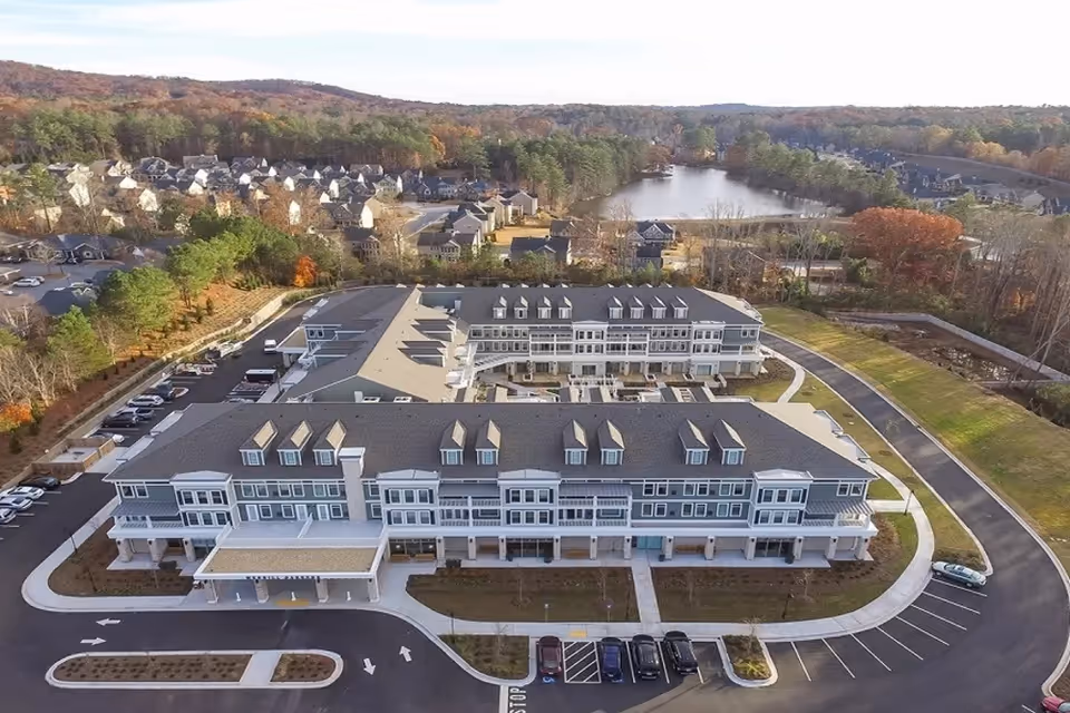 Aerial view of a large senior living facility building with multiple floors and dormer windows on the roof. The building is surrounded by parking lots, roads, and landscaped areas. In the background, there are residential houses, trees with autumn foliage, and a body of water.
