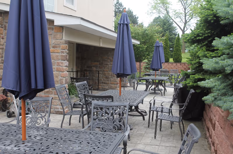 Outdoor patio area with multiple black metal tables and chairs, each table featuring a closed navy blue umbrella. The patio is adjacent to a building with stone and beige walls, surrounded by green trees and shrubs.