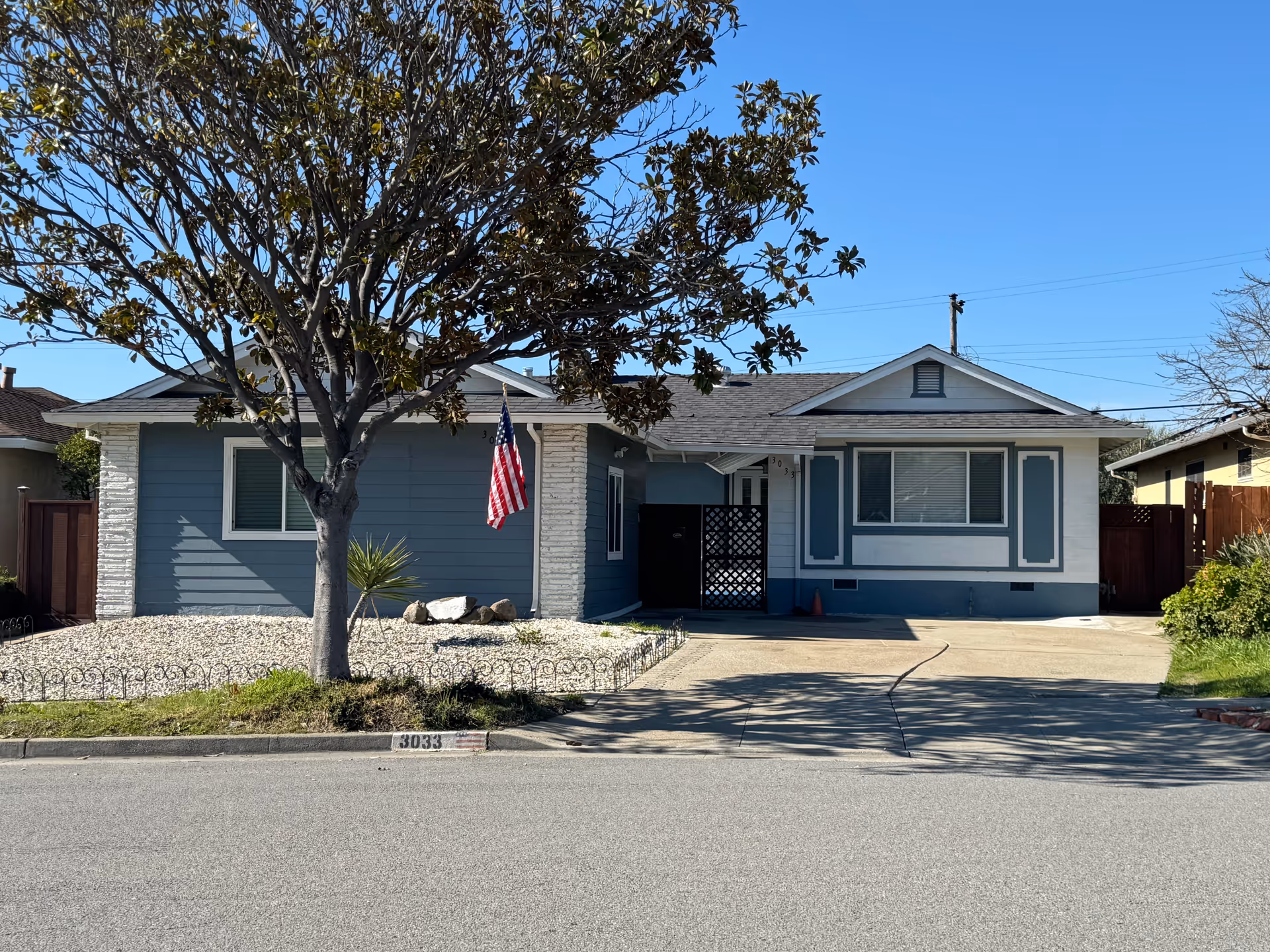 Single-story blue and white house with an American flag, a front tree, gravel landscaping, and a driveway under a clear blue sky.