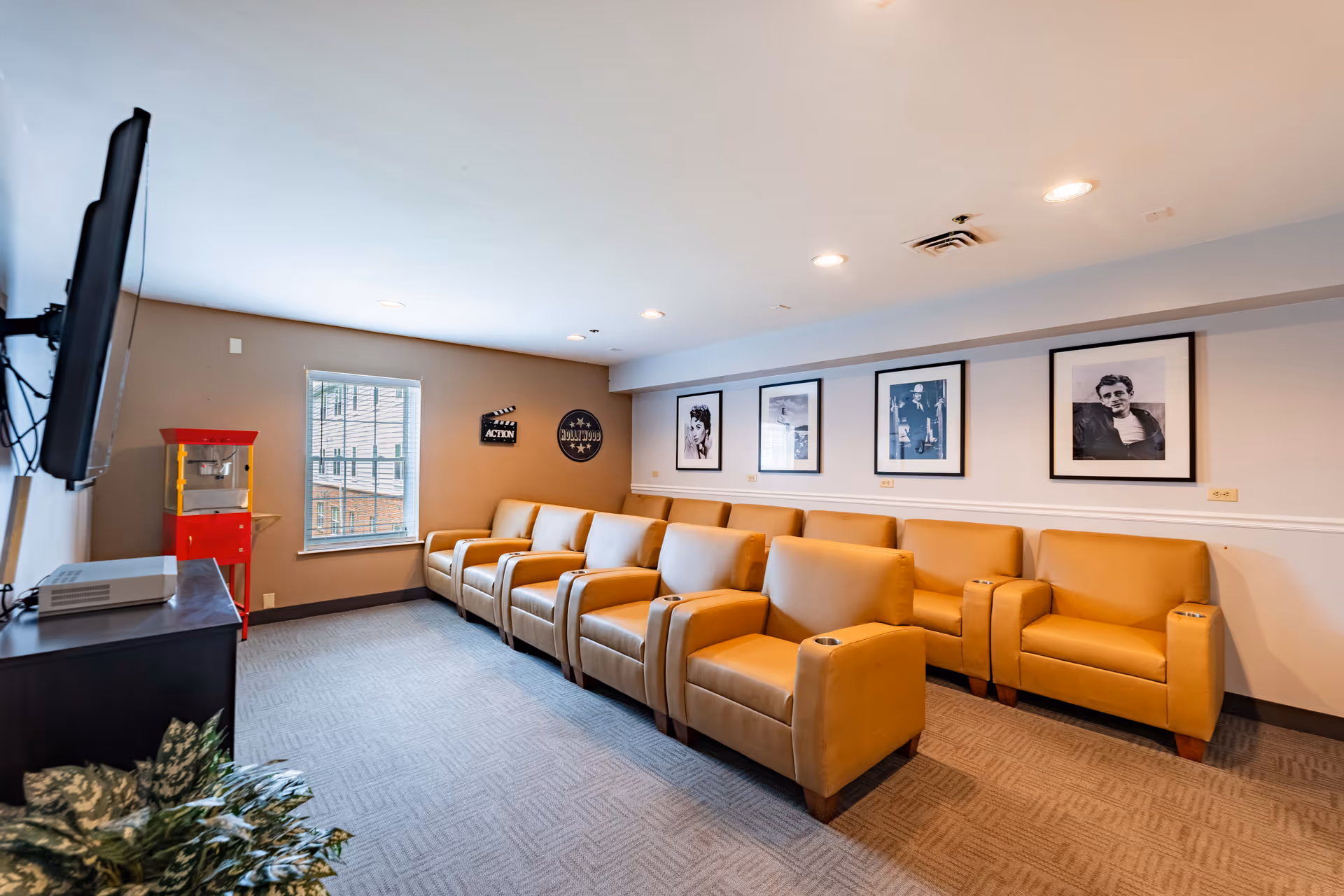 A small theater room with two rows of tan leather recliner chairs facing a wall-mounted flat screen TV. The room has beige walls with framed black and white photos of classic movie stars, a window with blinds, a red popcorn machine in the corner, and a carpeted floor.
