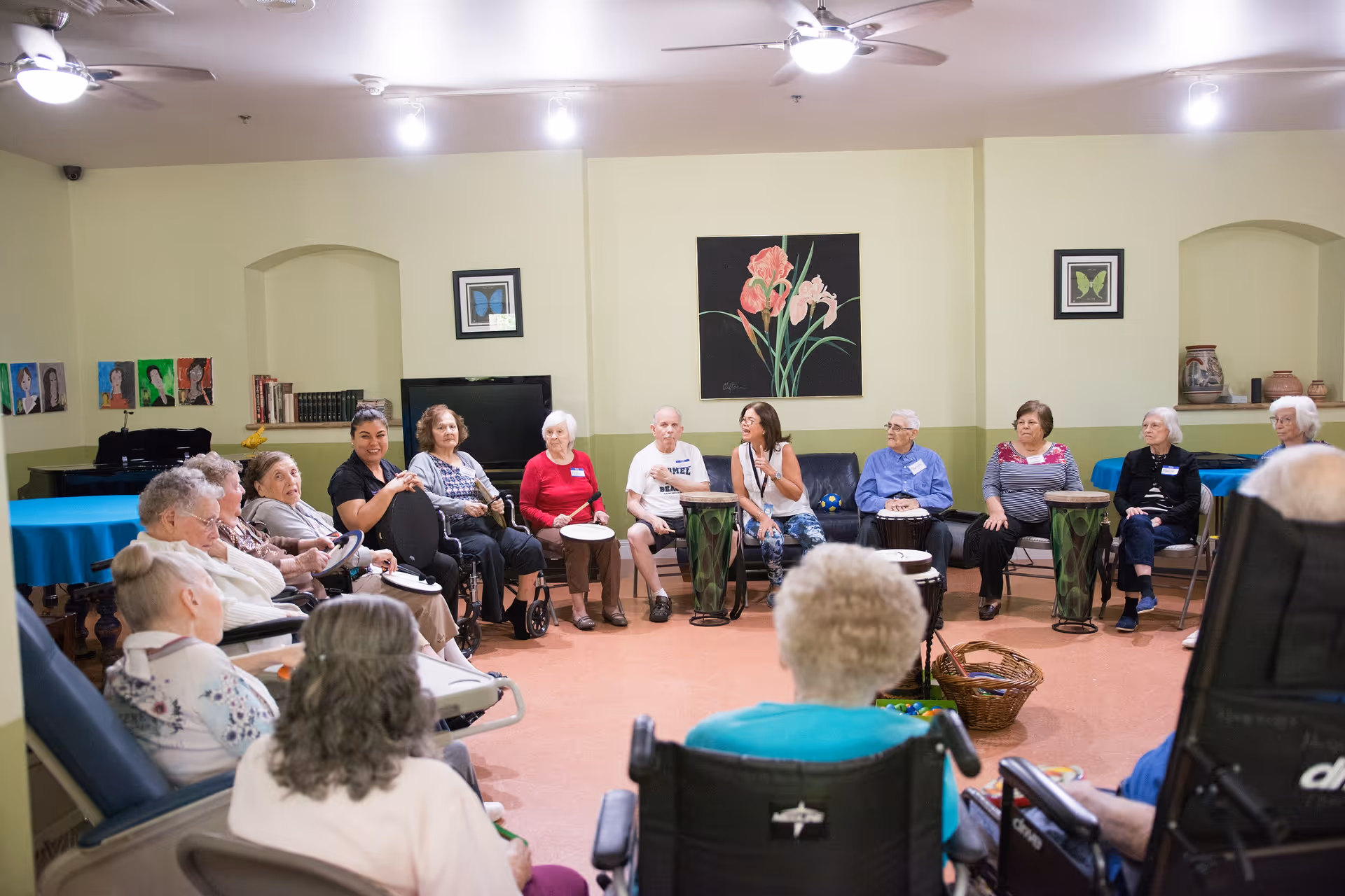 A group of elderly people sitting in a circle in a well-lit room, some in wheelchairs and others on chairs, participating in a drum circle activity with drums placed in front of them. The room has light yellow walls with framed artwork and a ceiling fan.